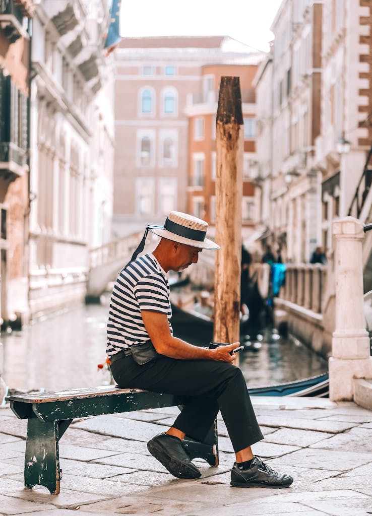 A gondolier in Venice relaxes on a bench by the canal, wearing a striped shirt and boater hat.