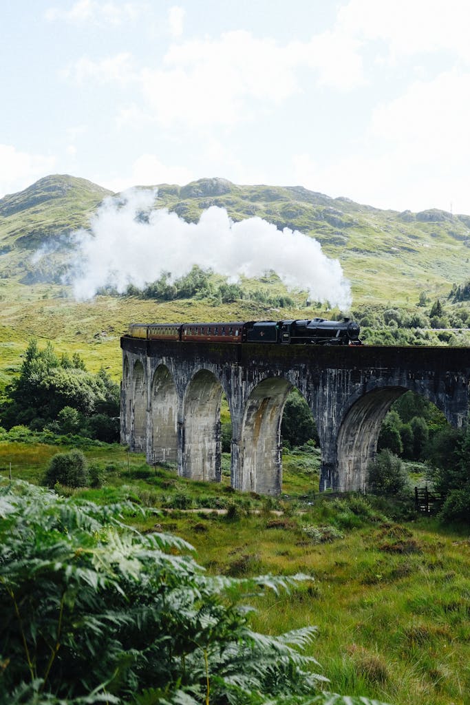 A scenic view of a steam train crossing the Glenfinnan Viaduct amidst lush green hills in Scotland.