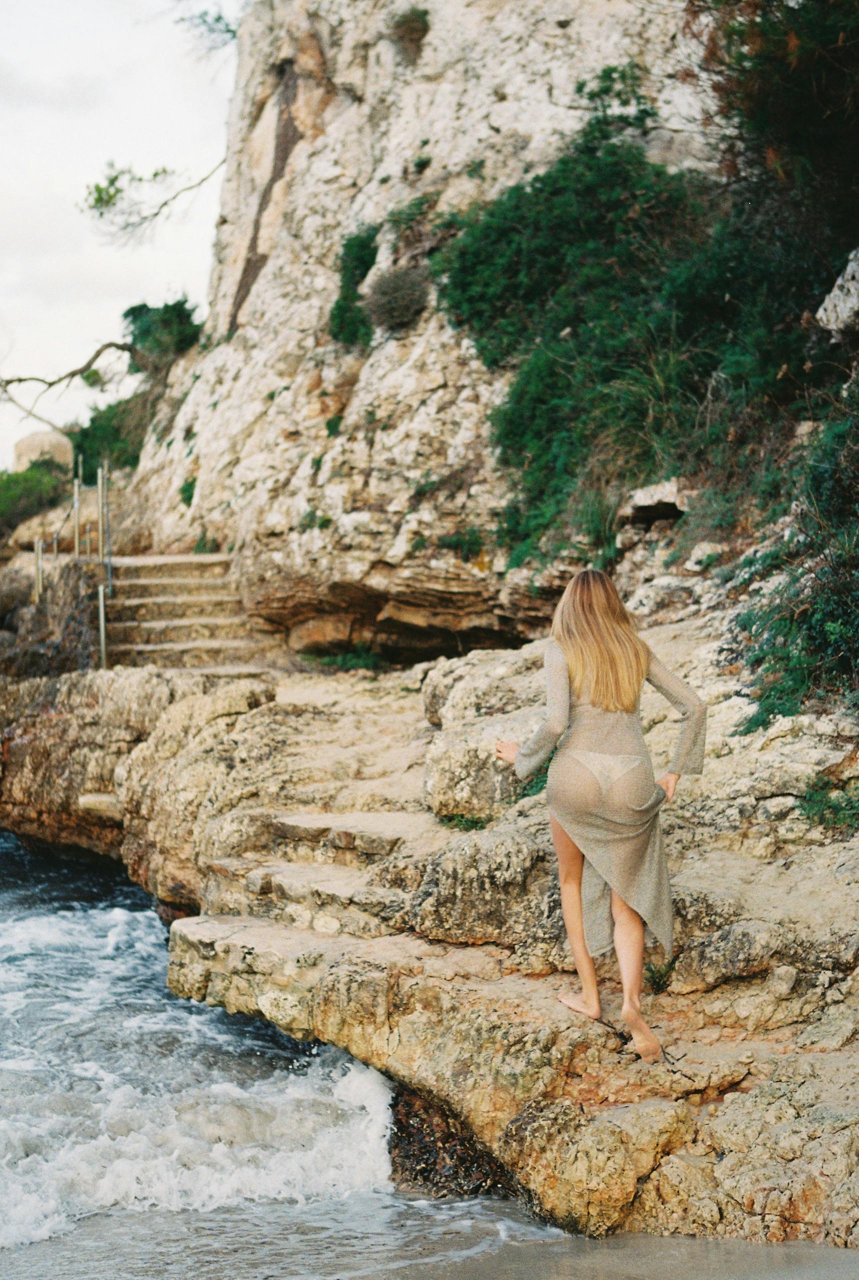 A woman in a dress walks barefoot along the rocky seashore of Cala Llombards, Spain.