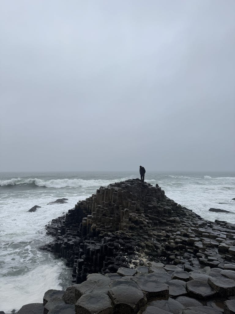 A lone figure stands on the iconic basalt columns of Giant's Causeway on a gray day.