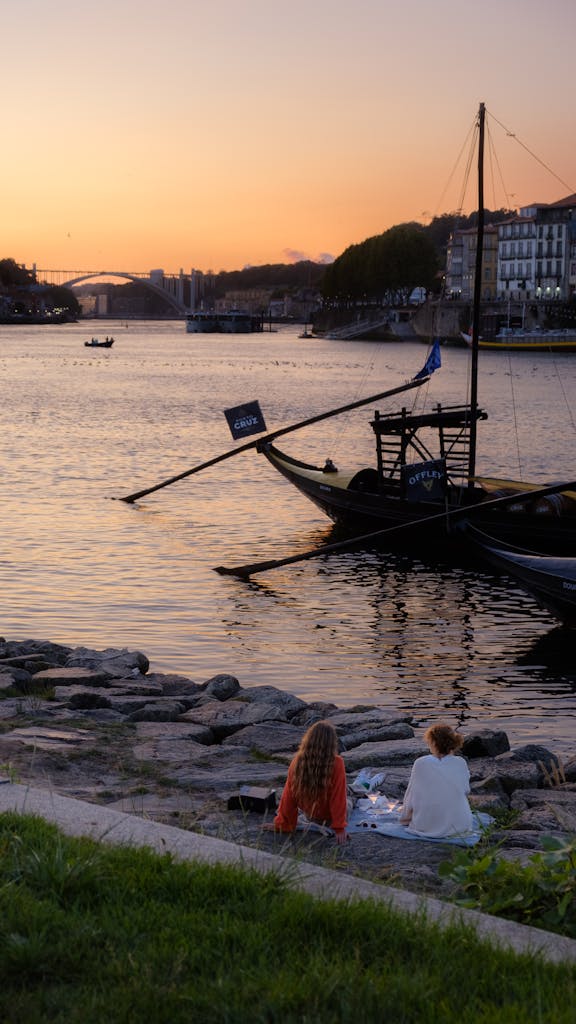 Two women enjoy a riverside sunset with traditional boats on the Douro River in Porto.